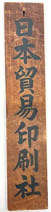 縦長の木の看板。墨で「日本貿易印刷社」と書かれている