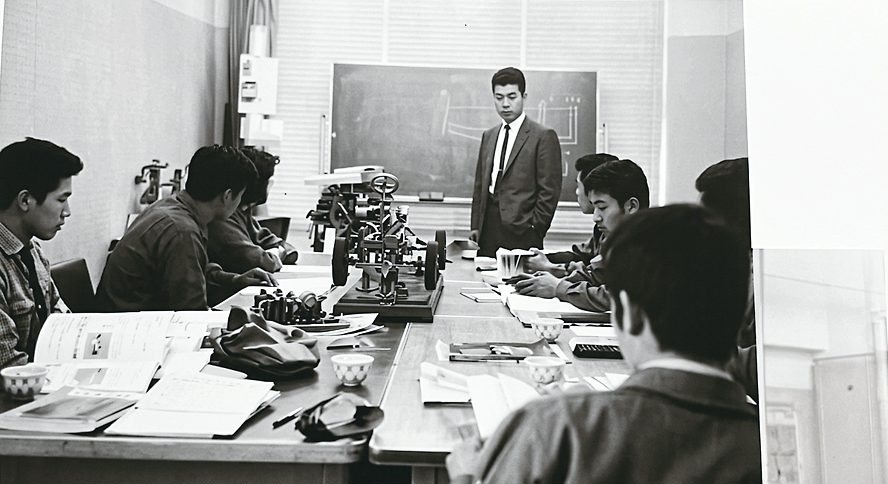 長机に座った受講者たちと、前に立つ講師。黒板のある研修室のモノクロ写真
