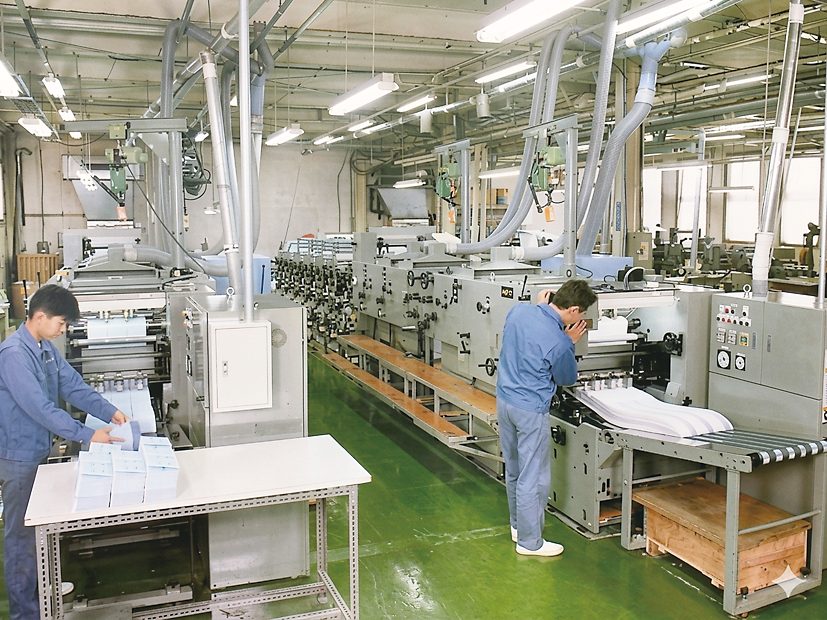 緑の床の工場で、大型の輪転印刷機を作業員2人が操作している