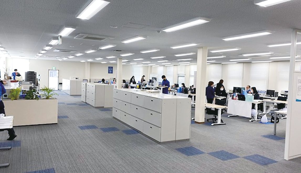 明るい執務フロア。低い収納棚が並び、奥で社員がパソコンに向かっている